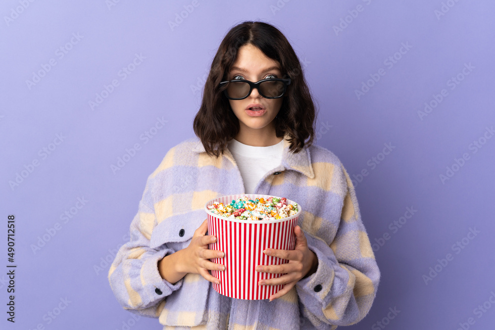 Teenager Ukrainian girl isolated on purple background surprised with 3d glasses and holding a big bucket of popcorns