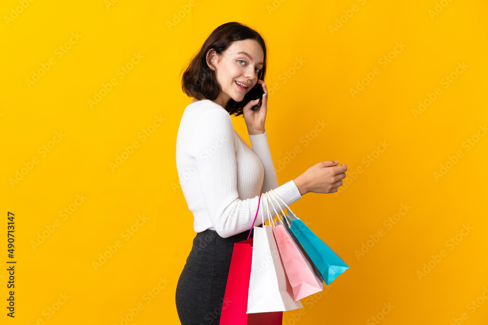 Teenager Ukrainian girl isolated on yellow background holding shopping bags and calling a friend with her cell phone Teenager Ukrainian girl isolated on yellow background holding shopping bags and calling a friend with her cell phone