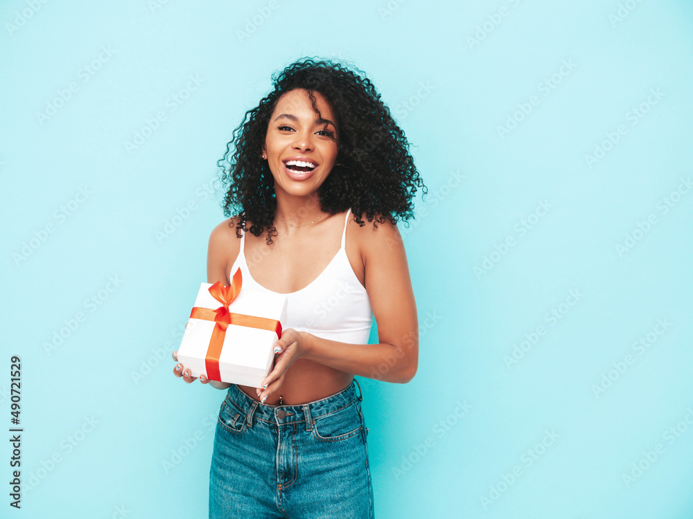 Beautiful black woman with afro curls hairstyle. Smiling model dressed in white summer clothes. Sexy carefree female posing near blue wall in studio. Tanned and cheerful. Holding gift box. Isolated