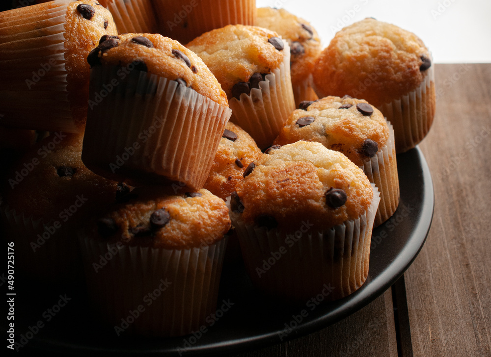 Close up or macro photography, vanilla cupcakes with chocolate chips on a white plate over a wooden lit by a window light. concept of homemade sweet bread. Horizontal Close up or macro photography, vanilla cupcakes with chocolate chips on a white plate over a wooden lit by a window light. concept of homemade sweet bread. Horizontal