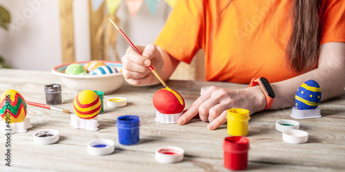 Woman's hand with a brush is drawing a colored pattern on an Easter egg. Creative preparation for bright Easter holiday