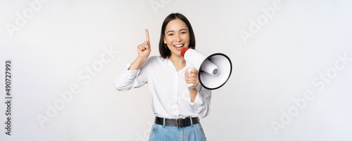 Smiling happy asian girl talking in megaphone and pointing up, announcing discount promo, showing advertisement on top, standing over white background photo