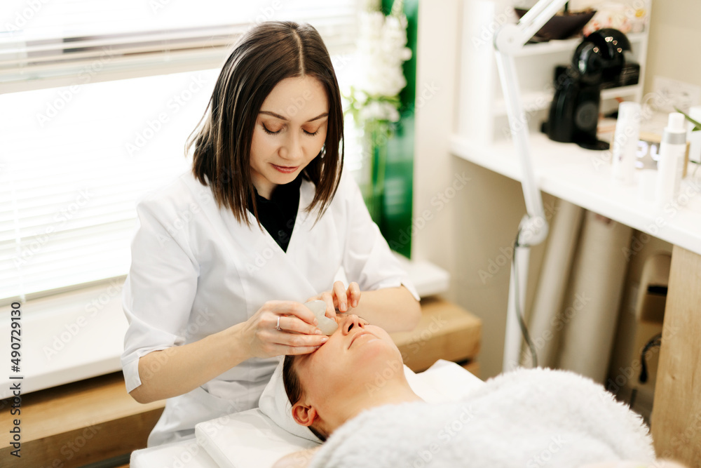 A cosmetologist massages the skin of a young woman with a gouache scraper. Facial massage, skin care, treatments A cosmetologist massages the skin of a young woman with a gouache scraper. Facial massage, skin care, treatments