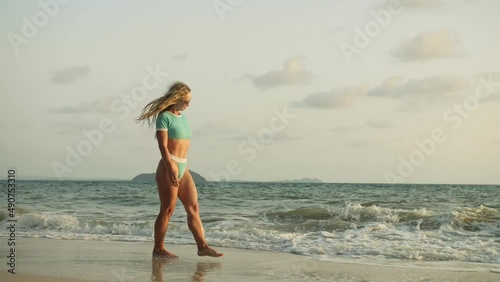 Sexy woman walking alone on beach coastline and relax warm golden sunset. Woman walking on water in blue swimsuit and sunglasses. Concept rest tropical resort traveling tourism happy summer holidays