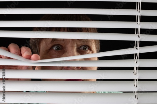 An elderly woman peers through the blinds through the window.