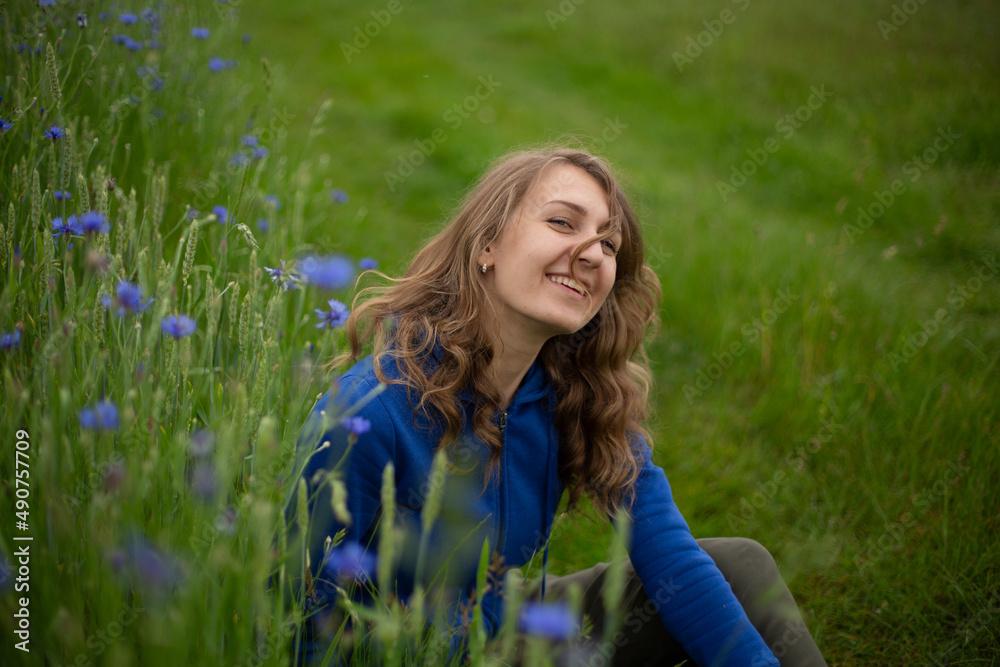 Satisfied blond girl is sitting on green grass near blue wildflowers. Women is wearing dark blue hoodie near cornflowers on the green background.
Satisfied blond girl is sitting on green grass near blue wildflowers. Women is wearing dark blue hoodie near cornflowers on the green background.