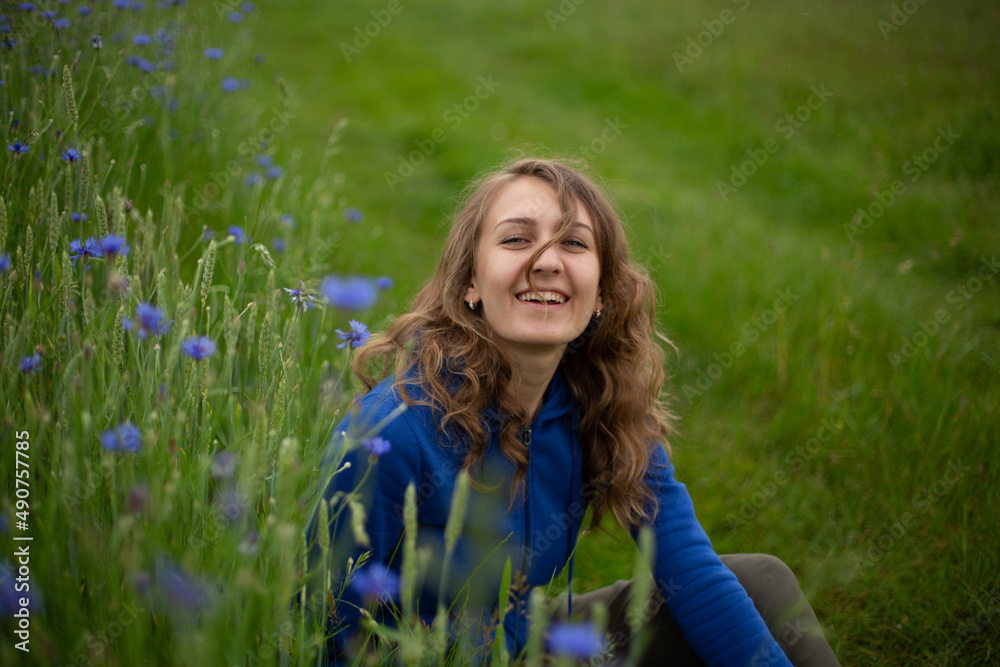 Satisfied blond girl is sitting on green grass near blue wildflowers. Women is wearing dark blue hoodie near cornflowers on the green background.
Satisfied blond girl is sitting on green grass near blue wildflowers. Women is wearing dark blue hoodie near cornflowers on the green background.