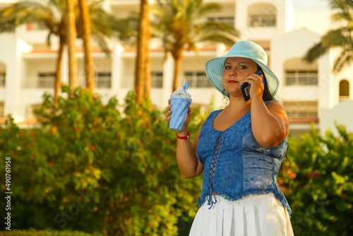 A young woman in a blue hat among palm trees and greenery against the background of the hotel speaks on a mobile phone.