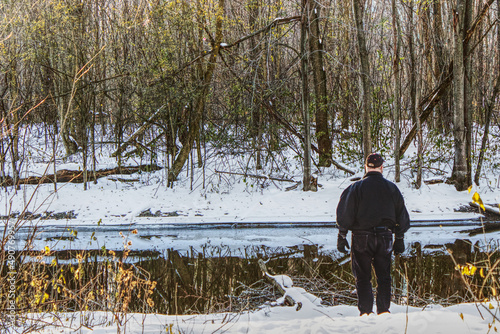 A man standing on the edge of the river, after the first snow, looking at the reflection of the trees in the water that is not yet frozen