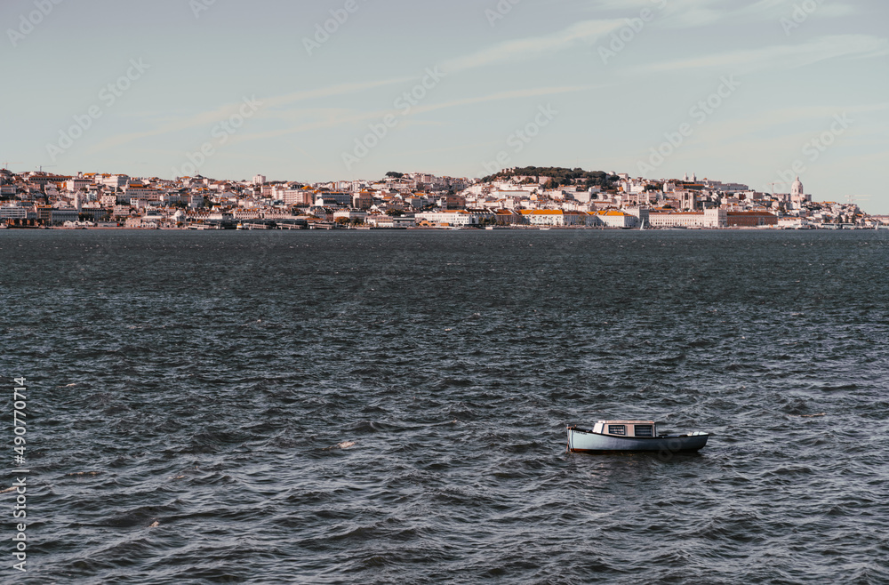An old tiny cozy boat with blue side plating and striped curtains inside of its cabin, sways in the waves of Tejo river, Portugal, with a riverbank full of small antique Lisbon houses in a background