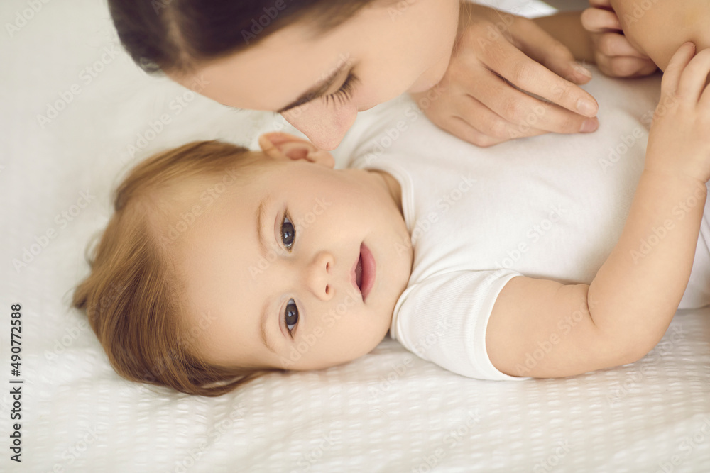 Little child together with mom, close up portrait. Happy beautiful young mother and her cute baby with soft fair hair cuddling on clean white sheets in comfy warm bed. Family, love, and care concept Little child together with mom, close up portrait. Happy beautiful young mother and her cute baby with soft fair hair cuddling on clean white sheets in comfy warm bed. Family, love, and care concept