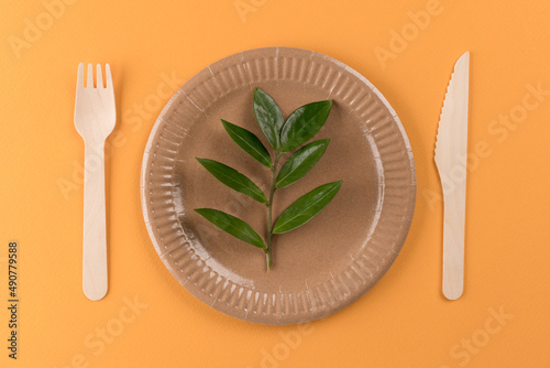 Eco-friendly tableware set. Kraft paper plate with wooden bamboo cutlery set on orange background and green leaves. Street food paper utensils. Ethical consumerism. Selective focus