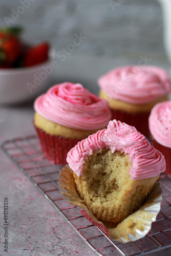 vanilla muffins decorated with pink buttercream frosting on top in red baking cups, on grey marble table and white stone wall