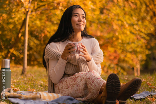 Portrait of cute girl sitting and having a cup of tea in a park picnic dressed fashionable on a October fall day.
