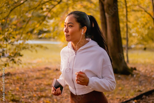 Slim asian fitness model jogging in a park with earbuds on a autumn day, living healthy running exercising in nature.