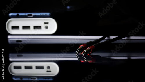 Red and black usb type-c connectors next to modern gadgets: laptop, an external battery, power bank and smartphone. Black background. Transferring data and charging gadgets. Selective focus