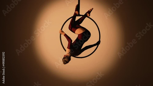 A pretty aerialist is spinning upside down on an aerial ring high under the dome of the circus. A young woman performs acrobatic stunts on a dark backlit studio background. Slow motion. photo