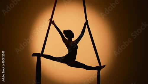 Silhouette of a young woman performs acrobatic elements and twine on airy silk. An aerial gymnast demonstrates stretching while performing a trick at height in a dark backlit studio. Slow motion. photo