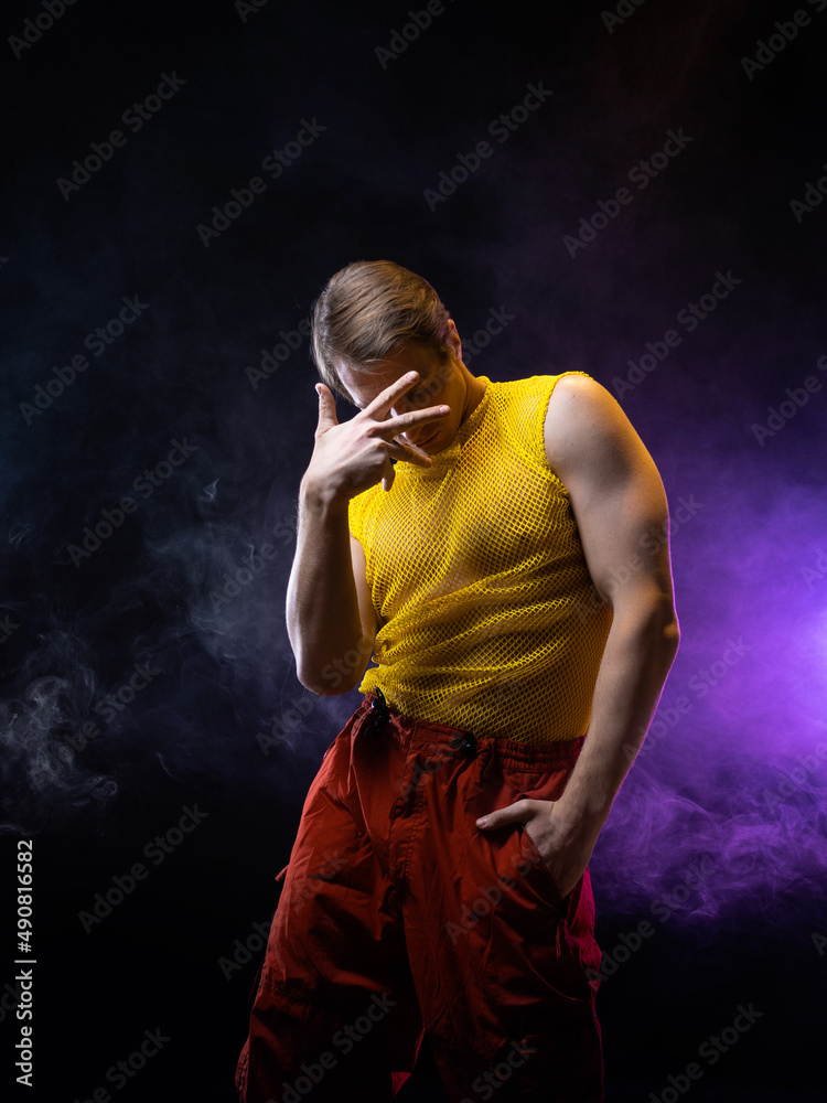 a young muscular man in a bright outfit poses in a pretentious pose, style and grotesque. Attractive guy with pumped-up arms in a mesh T-shirt and red pants, photo in smoke