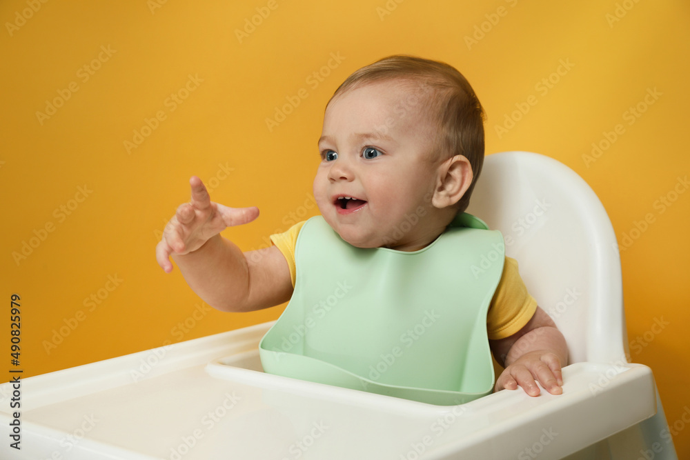 Cute little baby wearing bib in highchair on yellow background Cute little baby wearing bib in highchair on yellow background