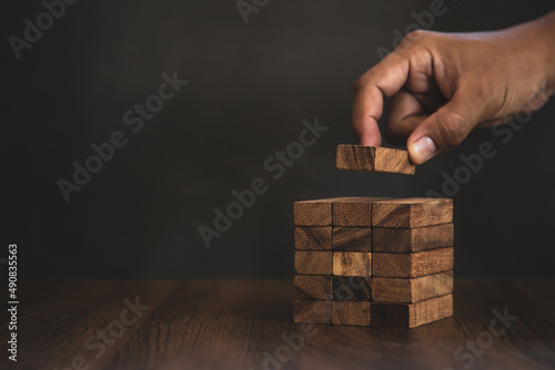 Close-up hand is placing wood block tower stacked with caution to prevent collapse or crash concepts of financial risk management and strategic planning.