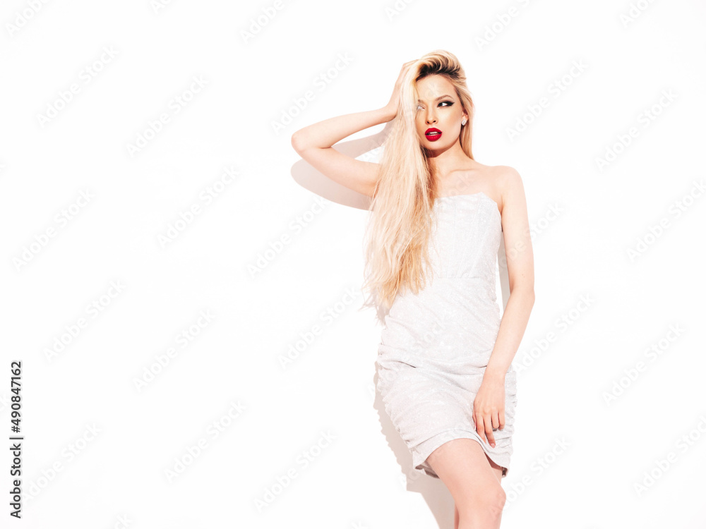 Young beautiful confident female in trendy summer evening silver dress. Sexy carefree blond  woman posing near white wall in studio. Model with red lips. Isolated