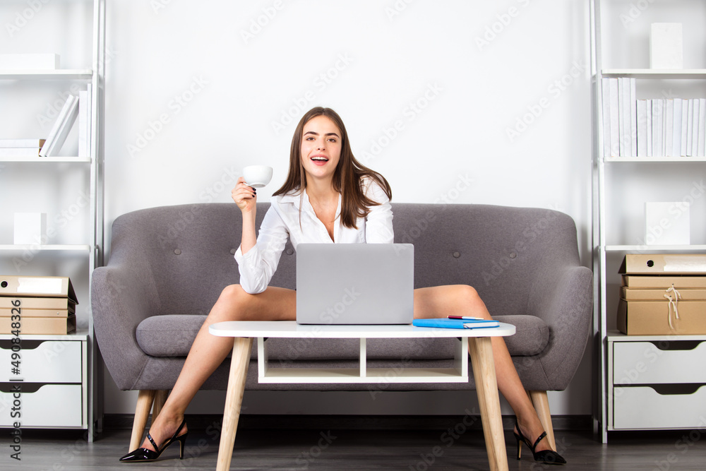 Attractive sexy business woman or secretary with laptop computer wearing  white suit with decollete sitting on sofa. Pretty accountant woman with cup  coffee at desk in office interior. Sexy long legs. Stock