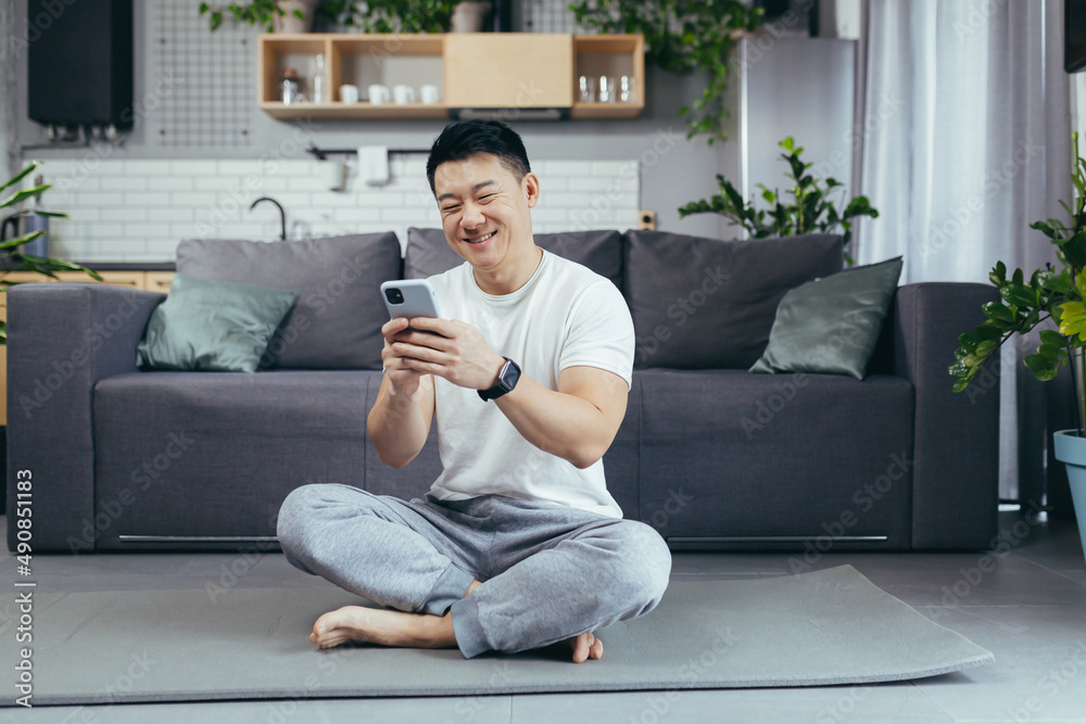 Obraz premium Male asian athlete at home using phone sitting on the floor during morning exercise, athlete in living room