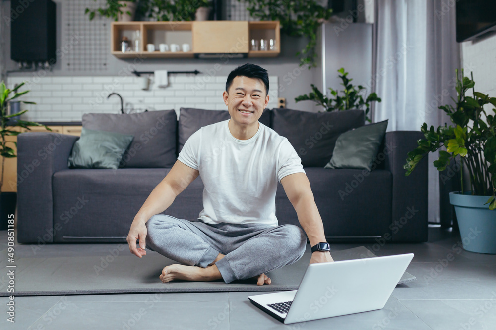 Happy Asian practices yoga at home, sitting in lotus position, man uses laptop for online classes, leads an active lifestyle Happy Asian practices yoga at home, sitting in lotus position, man uses laptop for online classes, leads an active lifestyle