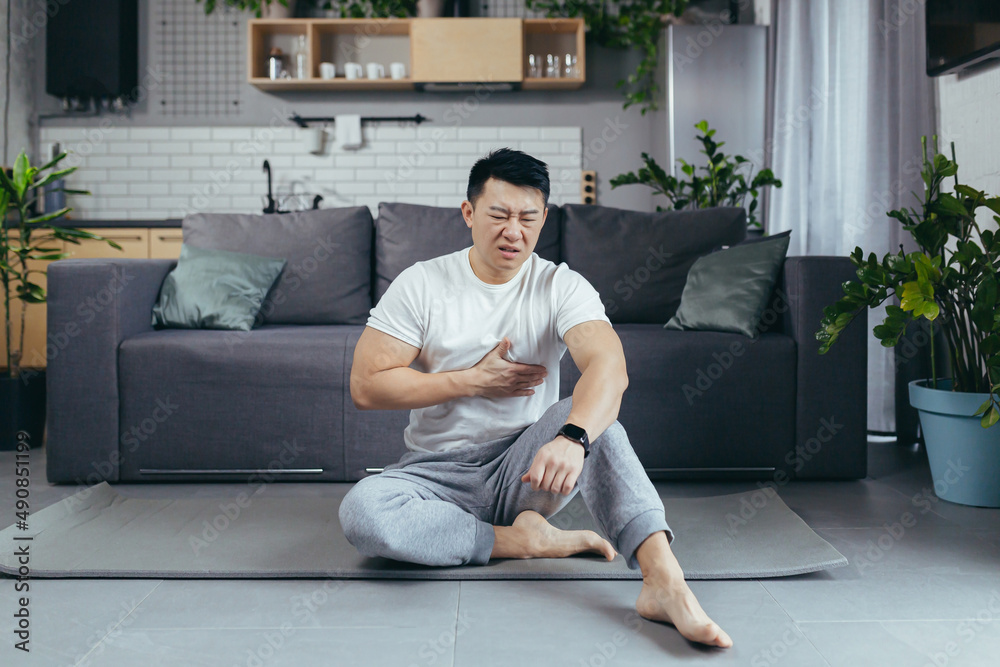 Athlete at home has severe heart pain after training, Asian sits on the floor and holds his hands to his chest Athlete at home has severe heart pain after training, Asian sits on the floor and holds his hands to his chest