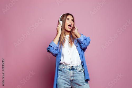 Beautiful young woman in headphones listening to music and singing on pink color background © artmim