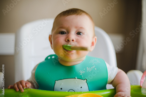 A baby in a bib is sitting on a high chair in front of the table, smiling, eating mashed potatoes with a spoon. Looks at the camera