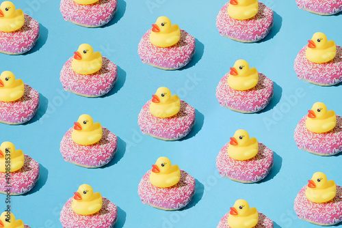 Small yellow rubber duck toy on donut with pink glaze and small white crumbs. Blue pastel background. Minimal design. Pattern.
