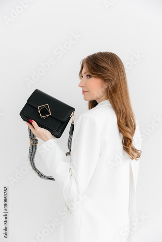 High fashion portrait of a young blonde model in a classic white suit. Vogue style. The girl sits at a table against a white background and holds a small leather handbag. 