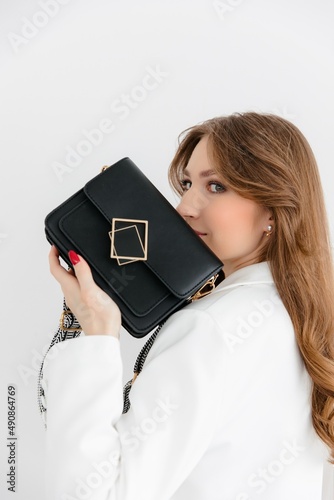 High fashion portrait of a young blonde model in a classic white suit. Vogue style. The girl sits at a table against a white background and holds a small leather handbag. 