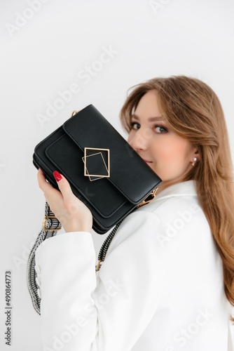 High fashion portrait of a young blonde model in a classic white suit. Vogue style. The girl sits at a table against a white background and holds a small leather handbag. 