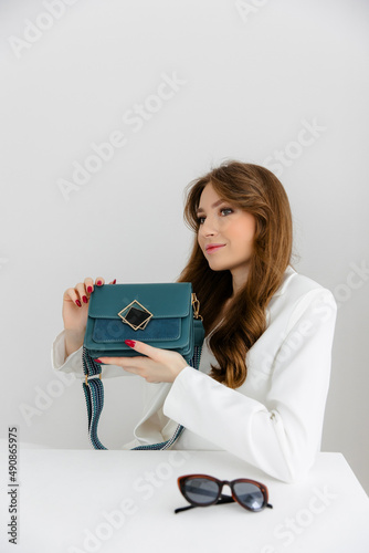 High fashion portrait of a young blonde model in a classic white suit. Vogue style. The girl sits at a table against a white background and holds a small leather handbag. 