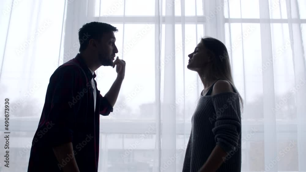 Married couple swears in living room background of curtains and windows, husband insults wife by pointing at her with finger, side view. Husband and wife quarrel at home. Concept of family problems