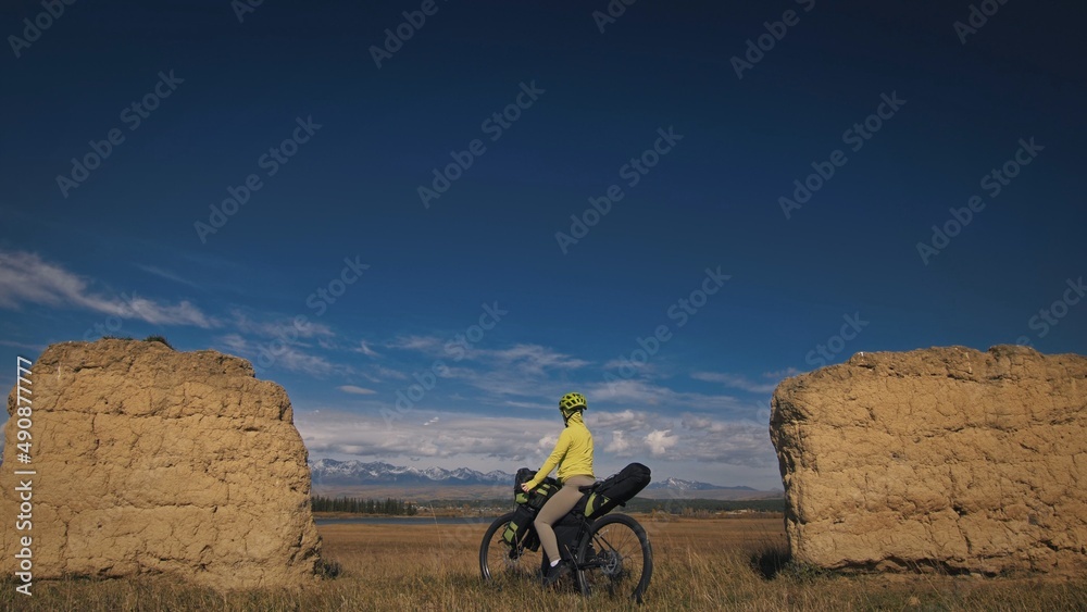 The woman travel on mixed terrain cycle touring with bikepacking. The traveler journey with bicycle bags. Sport bikepacking, bike, sportswear in green black colors. Mountain snow capped, stone arch.