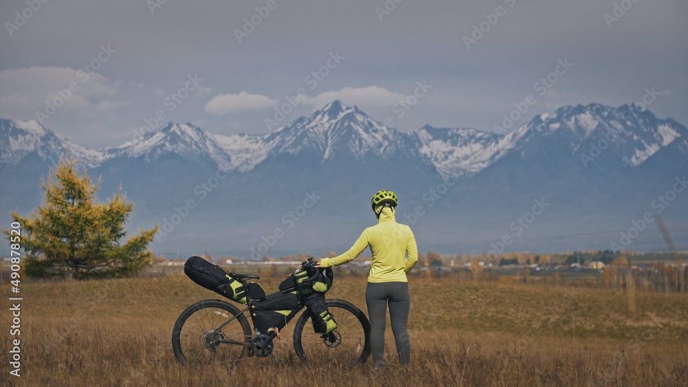 Obraz premium The woman travel on mixed terrain cycle touring with bikepacking. The traveler journey with bicycle bags. Sport tourism bikepacking, bike, sportswear in green black colors. Mountain snow capped.