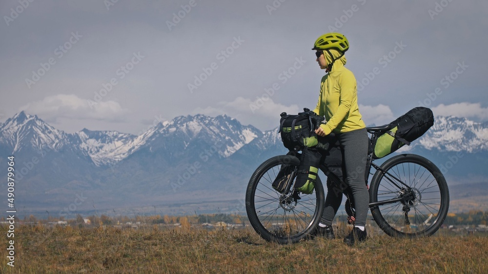 The woman travel on mixed terrain cycle touring with bikepacking. The traveler journey with bicycle bags. Sport tourism bikepacking, bike, sportswear in green black colors. Mountain snow capped. The woman travel on mixed terrain cycle touring with bikepacking. The traveler journey with bicycle bags. Sport tourism bikepacking, bike, sportswear in green black colors. Mountain snow capped.