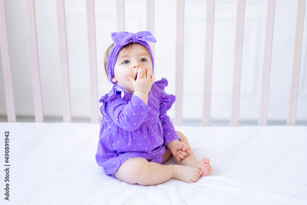 cute baby girl sitting on the bed in lilac clothes and with a bow on her head and smiling, funny little baby on a cotton bed at home playing, the concept of children's goods cute baby girl sitting on the bed in lilac clothes and with a bow on her head and smiling, funny little baby on a cotton bed at home playing, the concept of children's goods
