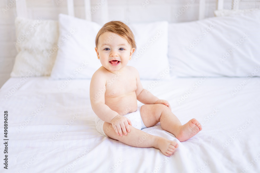 a joyful baby girl of six months on a bed in diapers smiles, a small child on a cotton bed at home, the concept of care and hygiene a joyful baby girl of six months on a bed in diapers smiles, a small child on a cotton bed at home, the concept of care and hygiene