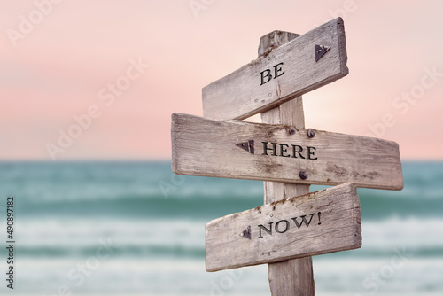 be here now text quote written on wooden signpost by the sea. Positive pink turqoise pastel theme.