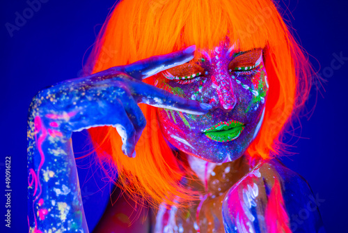 Close up UV portrait of a female model with artistic colorful makeup and orange wig, with closed eyes, isolated blue background.