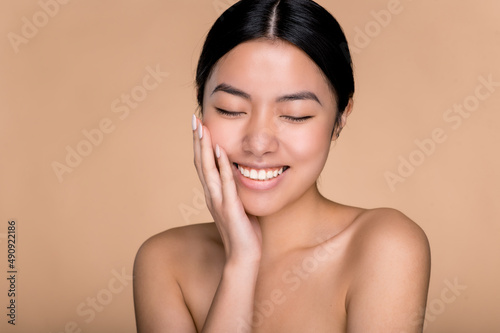 Beauty and care concept. A beautiful, happy Asian girl with perfect, well-groomed facial skin, with nude shoulders, stands on an isolated beige background, smiles with eyes closed