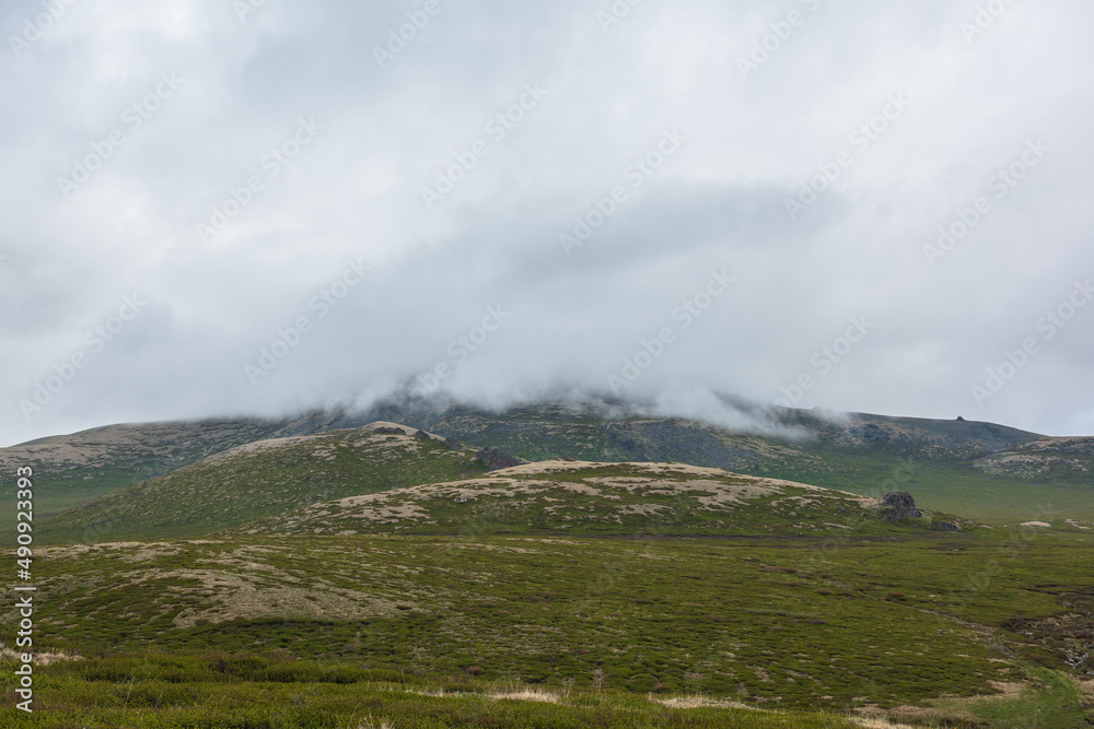 Dramatic alpine landscape with hills and mountains in gray low clouds. Bleak atmospheric scenery of tundra under lead gray sky. Gloomy minimalist view to green hills among low rainy clouds in overcast Dramatic alpine landscape with hills and mountains in gray low clouds. Bleak atmospheric scenery of tundra under lead gray sky. Gloomy minimalist view to green hills among low rainy clouds in overcast