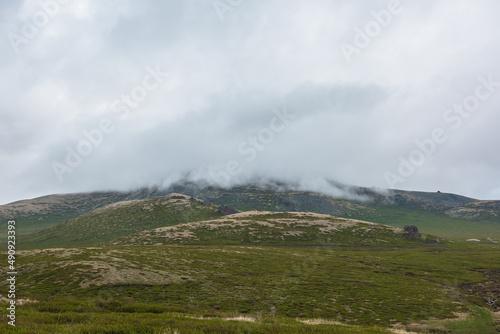 Dramatic alpine landscape with hills and mountains in gray low clouds. Bleak atmospheric scenery of tundra under lead gray sky. Gloomy minimalist view to green hills among low rainy clouds in overcast