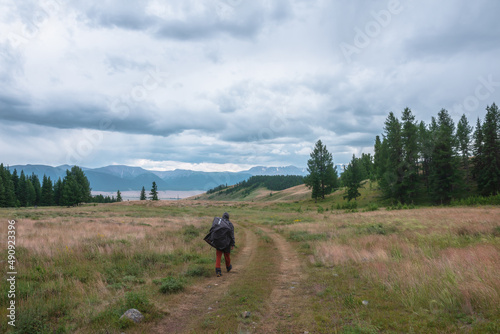 Tourist walks through hills and forest towards bad weather. Hiker on way to large snow mountain range under rainy cloudy sky. Man in raincoat in mountains in overcast. Traveler goes towards adventure. © Daniil