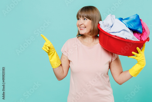 Elderly housewife woman 50s in t-shirt gloves hold clothes in basin basket after washing in washhouse point finger aside isolated on plain pastel light blue background Housekeeping tidying up concept photo